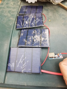 Vue de dessus de quatre mini panneaux solaires photovoltaïques bleus câblés en série par un fil rouge, reliés à un module convertisseur DC-DC boost avec sortie USB-A, posés sur un établi vert, avec un doigt pointant vers le module.