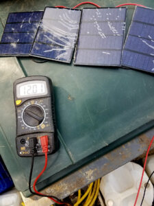 Quatre mini panneaux solaires photovoltaïques bleus câblés en série sur un établi vert, reliés à un multimètre numérique OHM affichant une tension de 12,01 volts en courant continu.