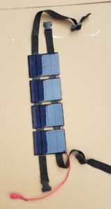 Chargeur solaire artisanal portable composé de quatre panneaux photovoltaïques bleus montés en colonne sur des sangles noires à boucles clip, avec un câble rouge terminé par un connecteur rose, posé sur un carton beige.