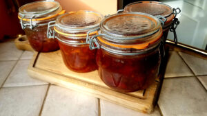 Quatre bocaux à joint orange en verre type Le Parfait, remplis d'un chutney ambré aux reflets brun-orangé, posés sur une planche à découper en bois, couvercles encore embués après stérilisation.