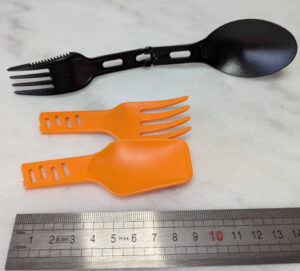 Deux options de couverts de randonnée posés sur une surface blanche avec une règle graduée : un couvert pliant noir combinant fourchette, couteau-scie et cuillère (environ 14 cm déplié), et un set de deux pièces orange en plastique rigide — une fourchette et une spatule-cuillère tronquées (environ 8 cm), séparables par un clip.