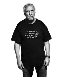 Portrait en noir et blanc de David Korn en t-shirt noir portant l'inscription humoristique « Go away or I will replace you with a very small shell script. »