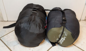 Deux sacs de couchage noirs compressés dans leur housse de rangement, posés côte à côte sur un carrelage blanc. Le sac de droite porte une étiquette verte de la marque Lafuma.