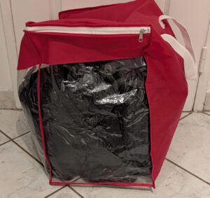 Sac de couchage noir rangé dans une grande housse de stockage rouge à fenêtre transparente et fermeture éclair blanche, posée sur un carrelage.
