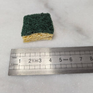 Petit carré d'éponge à récurer double face — mousse jaune et face abrasive verte — découpé à environ 4 cm de côté, posé sur une surface blanche à côté d'une règle graduée en centimètres.