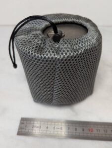Gamelle cylindrique en titane fermée par son couvercle, glissée dans une housse de protection en filet élastique gris à mailles larges fermée par un cordon noir, posée sur une surface blanche devant une règle graduée indiquant environ 10 cm de diamètre.