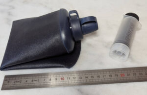 Gourde souple collapsible bleu marine à bouchon rotatif avec filtre intégré de marque LifeStraw, posée à plat sur une surface blanche à côté de sa seringue de rétro-lavage transparente et grise d'environ 10 cm, devant une règle graduée indiquant environ 15 cm de long pour la gourde à plat.
