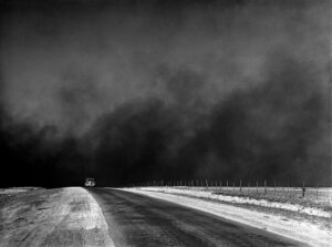 Photographie en noir et blanc d'un immense nuage de poussière obscurcissant le ciel au-dessus d'une route de terre rectiligne des Grandes Plaines, avec une automobile tentant de fuir la tempête, années 1930.