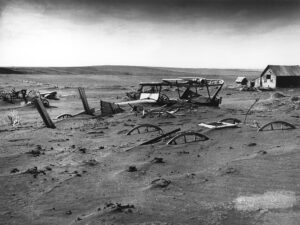 Photographie en noir et blanc d'une ferme abandonnée des Grandes Plaines, avec du matériel agricole — charrues, roues de chariots — à moitié enfoui sous des dunes de poussière et de sable, une grange en arrière-plan, sous un ciel menaçant, années 1930.