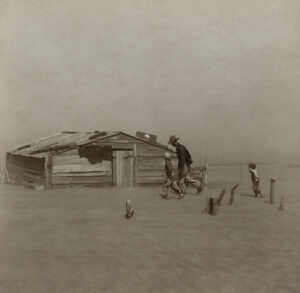 Photographie en sépia d'un homme courbé contre le vent et deux enfants se réfugiant contre une cabane en bois délabrée, battue par une tempête de poussière dans un paysage totalement désertique, Oklahoma, 1936.