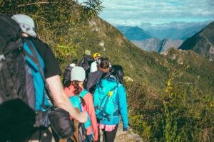 Groupe de randonneurs vus de dos, équipés de sacs à dos colorés, progressant en file indienne sur un sentier de crête étroit avec vue panoramique sur un massif montagneux verdoyant sous ciel nuageux.