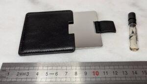 Deux objets posés sur une surface blanche devant une règle graduée : à gauche un miroir rectangulaire en métal poli argenté d'environ 8 cm glissé dans un étui en cuir noir cousu à languette de préhension, à droite un flacon échantillon en verre transparent d'environ 5 cm avec bouchon noir contenant un parfum ambré, portant une étiquette Paco Rabanne Invictus Glory.