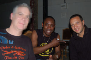 Trois hommes posant ensemble dans une salle, souriants et complices ; le visage de la personne de gauche est flouté.