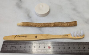 Trois objets posés sur une surface blanche devant une règle graduée : en haut un pastille de dentifrice solide blanc cylindrique d'environ 2 cm de diamètre, au centre un bâton de siwak brun naturel d'environ 14 cm aux fibres effilochées à une extrémité, en bas une brosse à dents en bambou gravée "Dentamyl" d'environ 17 cm aux poils blancs synthétiques.