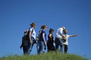 Groupe de six personnes en tenue décontractée marchant en file sur une crête herbeuse sous un ciel bleu vif, l'un d'eux pointant le doigt vers l'horizon, sans équipement technique particulier.