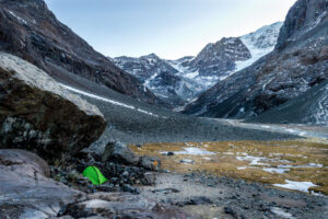 Petite tente de bivouac verte vif installée à l'abri d'un bloc rocheux au cœur d'un cirque glaciaire de haute montagne, entourée de parois rocheuses enneigées, d'éboulis et d'un torrent glaciaire, sous un ciel pâle d'altitude, sans présence humaine visible.