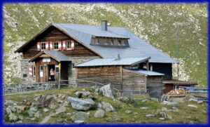 Grand bâtiment de refuge de montagne en bois et pierre, à toit métallique bleu, avec volets rouges et blancs, une terrasse avec tables et bancs, une annexe en bois, du bois coupé stocké à droite et deux personnes visibles à l'entrée, en contexte de pelouse alpine rocailleuse.