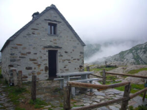 Petit bâtiment en pierre de taille à toit pentu, situé en terrain rocheux de haute montagne, avec une porte en bois ouverte, une fenêtre, une table et un banc en béton devant l'entrée, une barrière en rondins, dans un environnement brumeux et minéral.