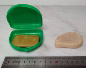 Deux produits d'hygiène posés sur une surface blanche devant une règle graduée : à gauche une petite boîte à protège-dents en plastique vert ouverte contenant un pain de savon jaune-ocre d'environ 5 cm, à droite un fragment de shampoing solide beige rosé en forme de galet d'environ 5 cm, tous deux posés devant une règle graduée.