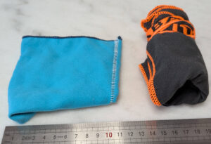 Deux articles de toilette en microfibre posés sur une surface blanche devant une règle graduée : à gauche un mini gant de toilette bleu turquoise plié en carré d'environ 10 cm, à droite une mini serviette en microfibre gris anthracite à liserés orange roulée sur elle-même d'environ 8 cm de diamètre.