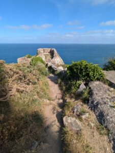 Sentier rocheux longeant une crête de falaise couverte de lande, menant vers des vestiges de maçonnerie en pierre dominant directement la Manche, avec des rochers de granite, de la végétation rase et un ciel bleu partiellement nuageux.