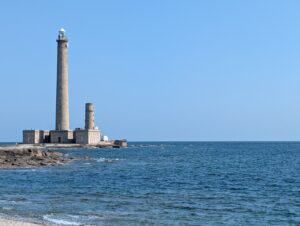 Vue depuis le rivage sur le phare de Gatteville et son phare secondaire, deux tours cylindriques en granite posées sur un ensemble de bâtiments bas au bord de la mer, sous un ciel bleu parfaitement dégagé, avec des rochers affleurants au premier plan.