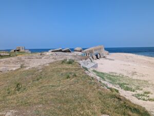 Anse sablonneuse bordée d'une lande herbeuse rase, avec plusieurs blocs de béton armé effondrés et inclinés — vestiges de blockhaus de la Seconde Guerre mondiale - éparpillés sur la plage et en bordure de dune, sous un ciel bleu sans nuages.
