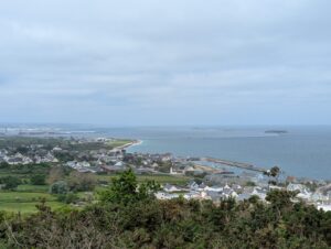 Vue panoramique en hauteur depuis la batterie de Bretteville-le-Haut sur l'agglomération de Tourlaville et la grande rade de Cherbourg, avec la digue du large, le fort de l'Île Pelée et les installations portuaires visibles à l'horizon, sous un ciel couvert et gris.