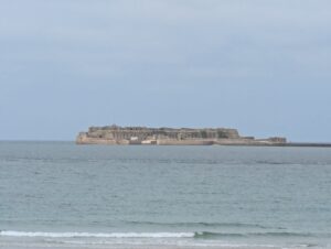 Vue depuis le rivage sur le fort de l'Île Pelée, vaste ensemble fortifié en pierre posé à fleur d'eau au milieu de la rade de Cherbourg, sous un ciel gris uniforme, avec une mer légèrement agitée au premier plan.