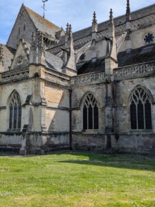 Vue rapprochée du flanc extérieur de la basilique Sainte-Trinité de Cherbourg-en-Cotentin, montrant des fenêtres à remplage gothique flamboyant, des pinacles, des gargouilles et une frise ajourée en pierre calcaire, avec une pelouse fleurie de marguerites au premier plan, sous un ciel bleu.