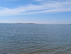 Vue depuis le rivage de Saint-Vaast-la-Hougue sur la Manche calme, avec l'île de Tatihou visible à l'horizon, sous un ciel bleu partiellement nuageux.