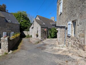 Ruelle pavée d'un petit hameau normand, bordée de maisons et murets en pierre de granite appareillée, avec des fleurs en pot et des volets blancs, sous un ciel bleu sans nuages.