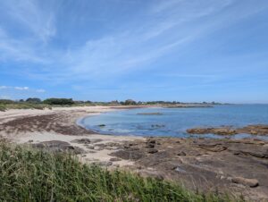 Vue panoramique depuis la pointe Maigret sur une anse sablonneuse bordée de rochers de granite, avec des eaux peu profondes aux reflets turquoise, des dunes herbeuses et quelques maisons à l'arrière-plan, sous un ciel bleu strié de nuages légers.