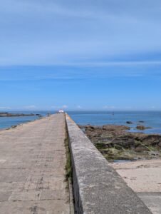Vue en perspective le long d'une jetée en pierre bétonnée s'avançant dans la mer, avec un petit feu de signalisation rouge et blanc à son extrémité, des rochers affleurant à droite et la Manche ouverte à l'horizon, sous un ciel bleu.