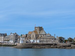 Vue depuis le port sur le bourg de Barfleur, dominé par l'église Saint-Nicolas en granite, entourée de maisons traditionnelles normandes, avec le quai et les rochers au premier plan et un ciel bleu dégagé.