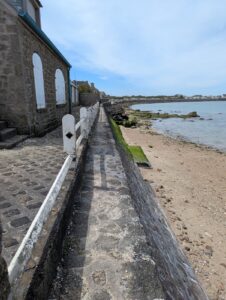 Passage piéton étroit en pavés longeant des maisons en granite à gauche et un muret surplombant une plage de sable et de rochers à droite, avec la baie de Barfleur et des habitations visibles à l'horizon, sous un ciel partiellement nuageux.