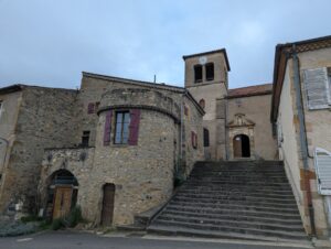 Place centrale de Saint-Maurice-ès-Allier par temps couvert, avec au premier plan un bâtiment en pierre de lave à tour cylindrique et volets bordeaux, et en arrière-plan l'église romane surmontée de son clocher-tour à horloge, accessible par un large escalier en pierre.