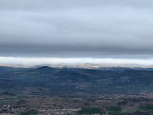 Vue panoramique depuis le sommet du Puy Saint-Romain sur un vaste paysage de collines auvergnates étagées, avec des zones urbanisées visibles dans la plaine et sur les versants, sous un ciel chargé de nuages gris laissant filtrer quelques trouées lumineuses à l'horizon.