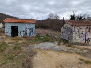 Deux petits bâtiments aux toits de tuiles romaines abritant des sources d'eaux salées sur sol sableux humide ; le bâtiment de gauche est orné d'un street art représentant fleurs et pichet versant de l'eau sur fond bleu, celui de droite est recouvert de tags multicolores. À l'arrière-plan, une ripisylve aux arbres dénudés sous ciel nuageux.