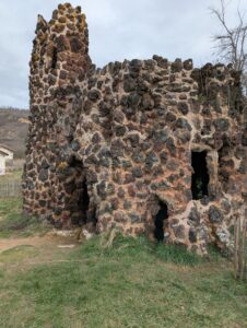 Construction de style faussement médiéval édifiée en pierres volcaniques sombres et mortier ocre rosé, percée de plusieurs ouvertures en arc irrégulier évoquant des grottes, surmontée d'une tourelle à sommet dentelé couverte de lichens jaunes, sur fond de coteau boisé et ciel nuageux.
