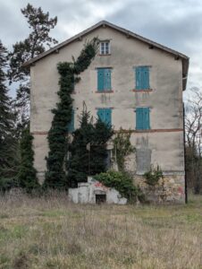 Bâtiment de trois étages à façade enduite beige, aux volets bleus délavés et partiellement fermés, envahi par un lierre dense grimpant jusqu'au pignon ; l'entrée principale est condamnée, le rez-de-chaussée partiellement muré, et le terrain environnant laissé à l'état de friche herbacée sous ciel nuageux.