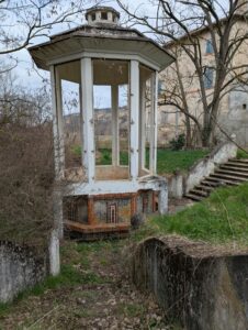 Kiosque octogonal à colonnes blanches et toiture en pavillon de béton, reposant sur un soubassement carrelé de mosaïques géométriques brun et ocre, abritant un bassin vide ; à droite, un escalier en pierre mène vers l'ancienne station thermale visible à l'arrière-plan, entourée d'arbres dénudés sous ciel nuageux.