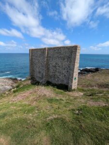 Mur épais en maçonnerie de moellons de granite, de forme rectangulaire à contreforts latéraux, dressé isolément sur un promontoire herbeux dominant directement la mer et des rochers, sous un ciel bleu à nuages blancs.