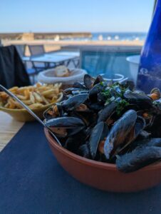 Bol en terre cuite rempli de moules marinières persillées, accompagné d'un plat de frites maison et d'une corbeille de pain, servis en terrasse face à la mer au crépuscule, le port d'Omonville-la-Rogue visible en arrière-plan.