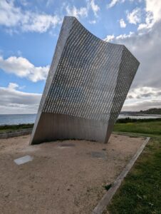 Grande sculpture monumentale en acier inoxydable perforé de texte en français, à la forme trapézoïdale légèrement incurvée, installée sur un espace de sable face à la mer, sous un ciel partiellement nuageux, sur la côte de Cherbourg-en-Cotentin.