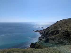 Vue en plongée depuis le sommet d'une falaise couverte de lande sur une mer d'un bleu profond aux reflets argentés, avec une paroi rocheuse sombre à droite sur laquelle se devine un sentier, des rochers affleurants en contrebas, sous un ciel bleu parfaitement dégagé.