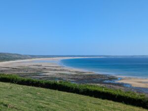 Vue en hauteur depuis une pelouse sur une vaste baie sablonneuse déserte en courbe ample, avec un estran découvert à marée basse présentant des flaques et des algues au premier plan, une mer turquoise et bleue, et des dunes bordant le rivage à l'horizon, sous un ciel bleu parfaitement dégagé.
