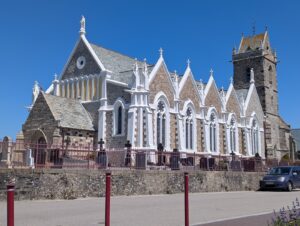 Façade latérale de l'église Saint-Pierre-et-Saint-Paul de Biville, édifice en granite appareillé aux encadrements gothiques peints en blanc, avec pinacles, rosaces et fenêtres à remplage, entourée d'un cimetière clos d'une grille bordeaux, sous un ciel bleu vif.