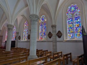Vue intérieure de la nef latérale de l'église Saint-Pierre-et-Saint-Paul de Biville, avec des colonnes en granite blanc soutenant des voûtes en ogive, une série de vitraux figuratifs multicolores à dominantes bleues et rouges, des médaillons de chemin de croix entre les fenêtres, et des bancs en bois clair au premier plan.