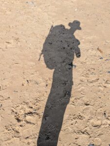 Ombre portée allongée d'un randonneur coiffé d'un chapeau et portant un sac à dos avec des bâtons de marche, projetée sur le sable humide et marqué de traces de pas de la plage de Siouville-Hague, en plein soleil.